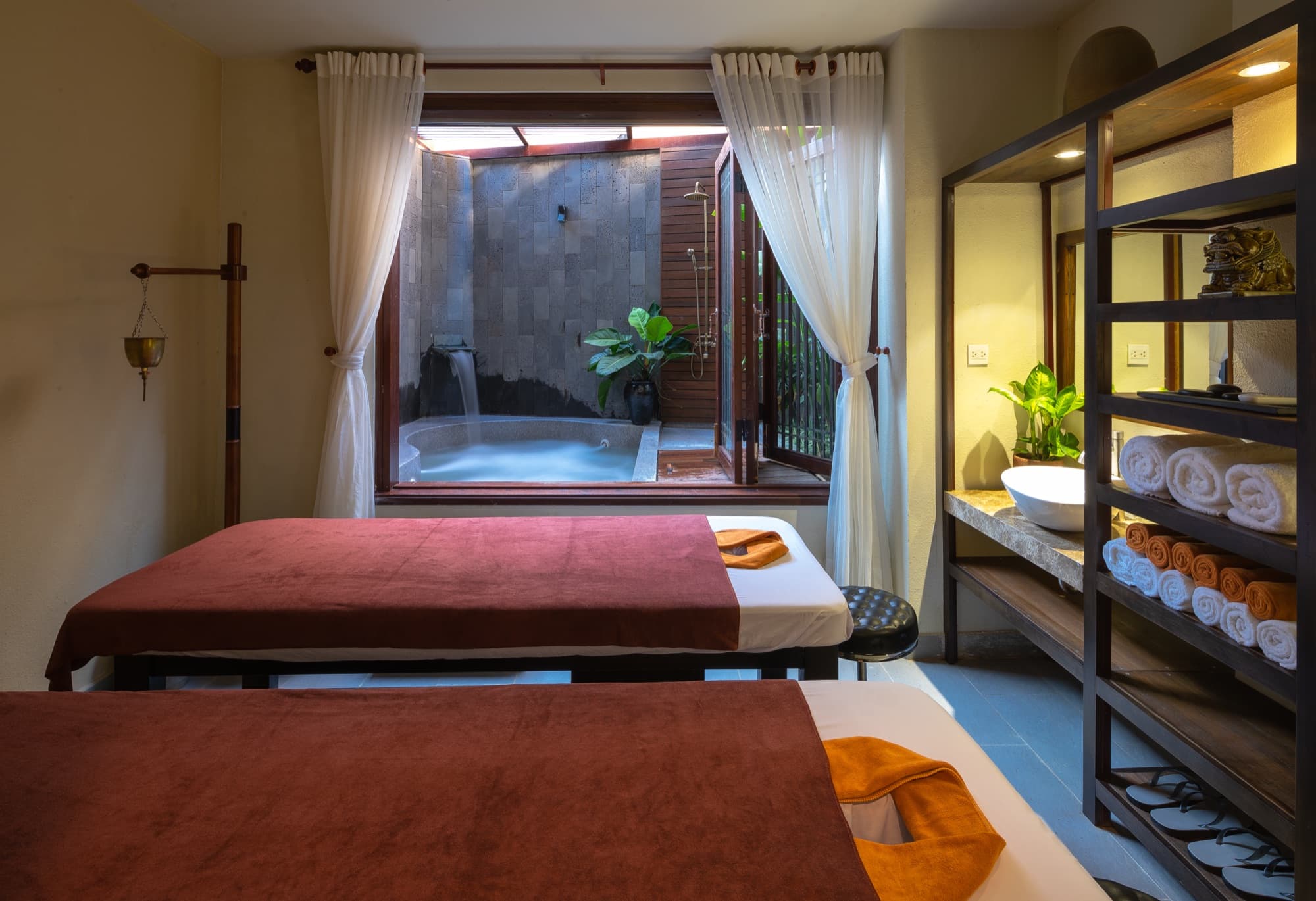 The private couple's treatment room at Nghê Prana, Hội An — two side-by-side beds and a private jacuzzi through the glass door, on the Thu Bồn at Cẩm Nam.