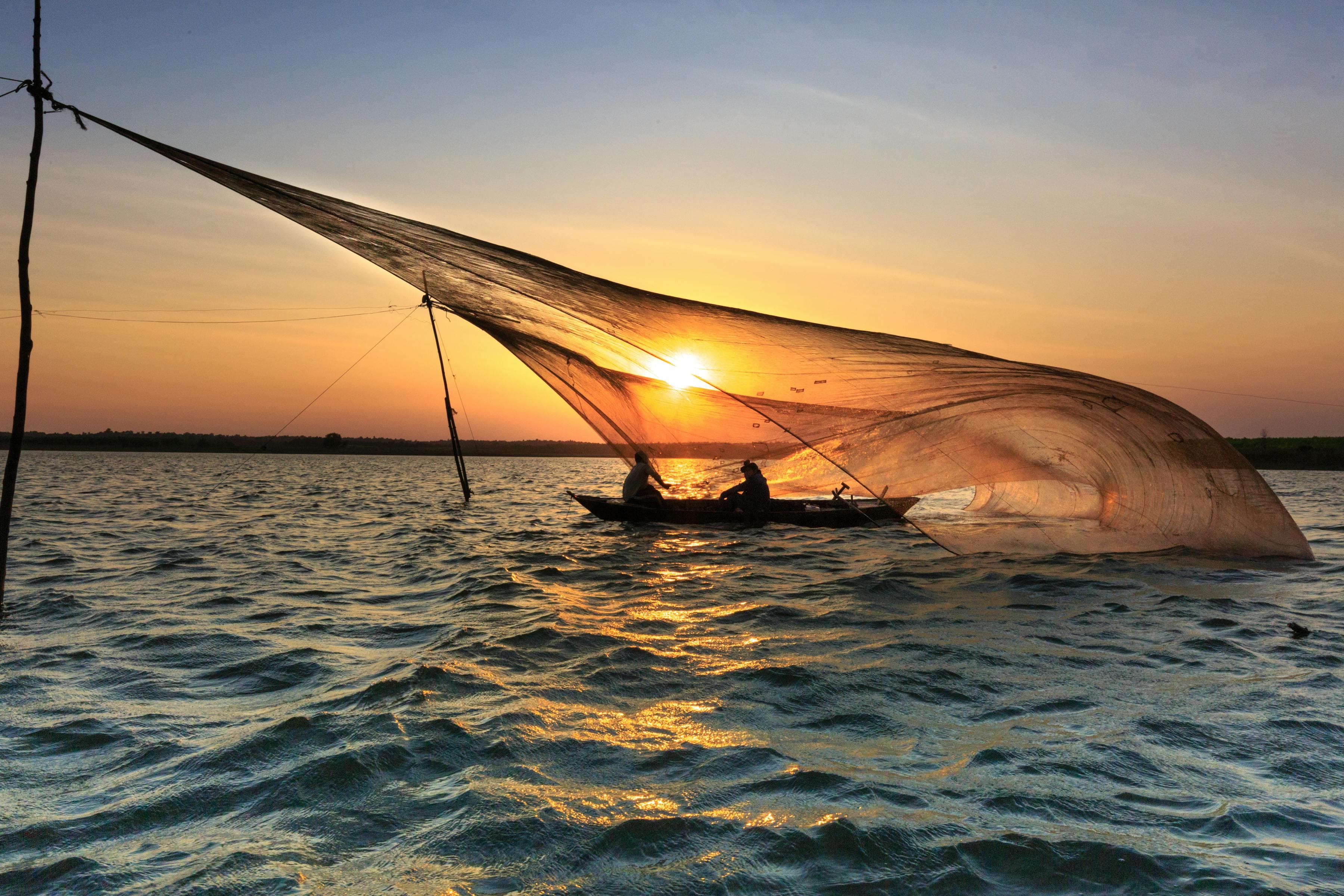 Vietnamese fishermen with a traditional square-net (rớ) silhouetted against a low golden sun over calm river water at dawn — sunrise on the Thu Bồn at Cửa Đại estuary, Hội An